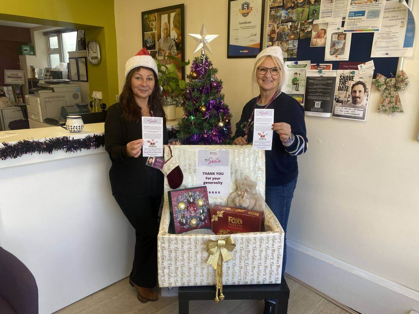 Two women in Santa hats holding flyers stand beside a decorated Christmas tree and a gift box filled with holiday items. - Home Instead