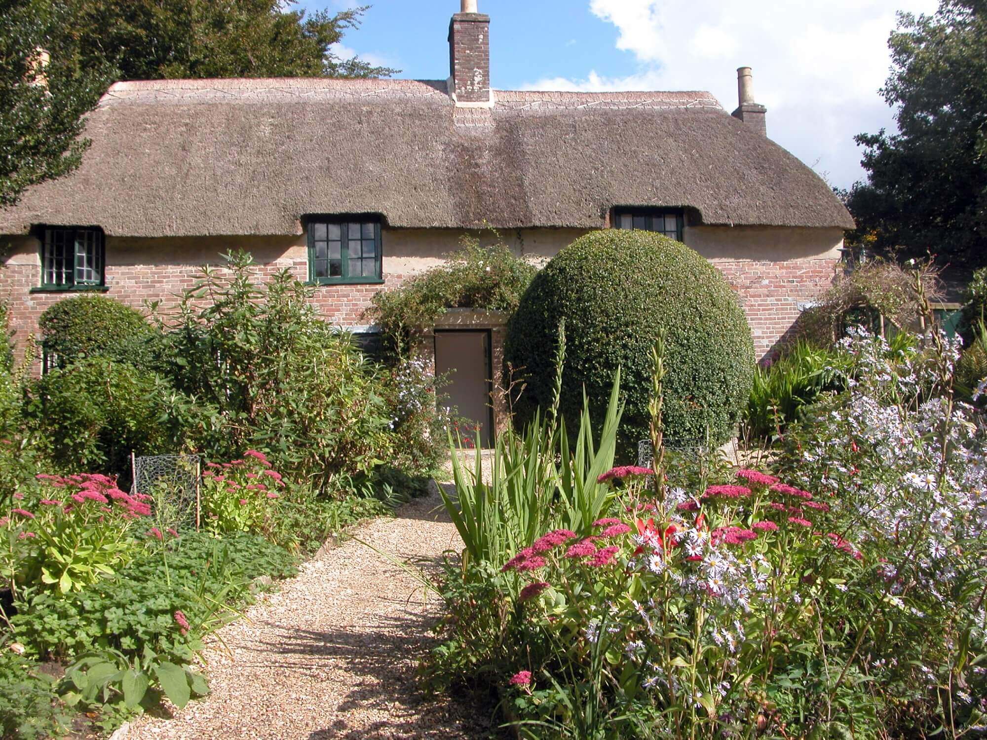 A quaint cottage with a thatched roof surrounded by a lush, colourful garden and a gravel path leading to the door. - Home Instead Bournemouth & Christchurch