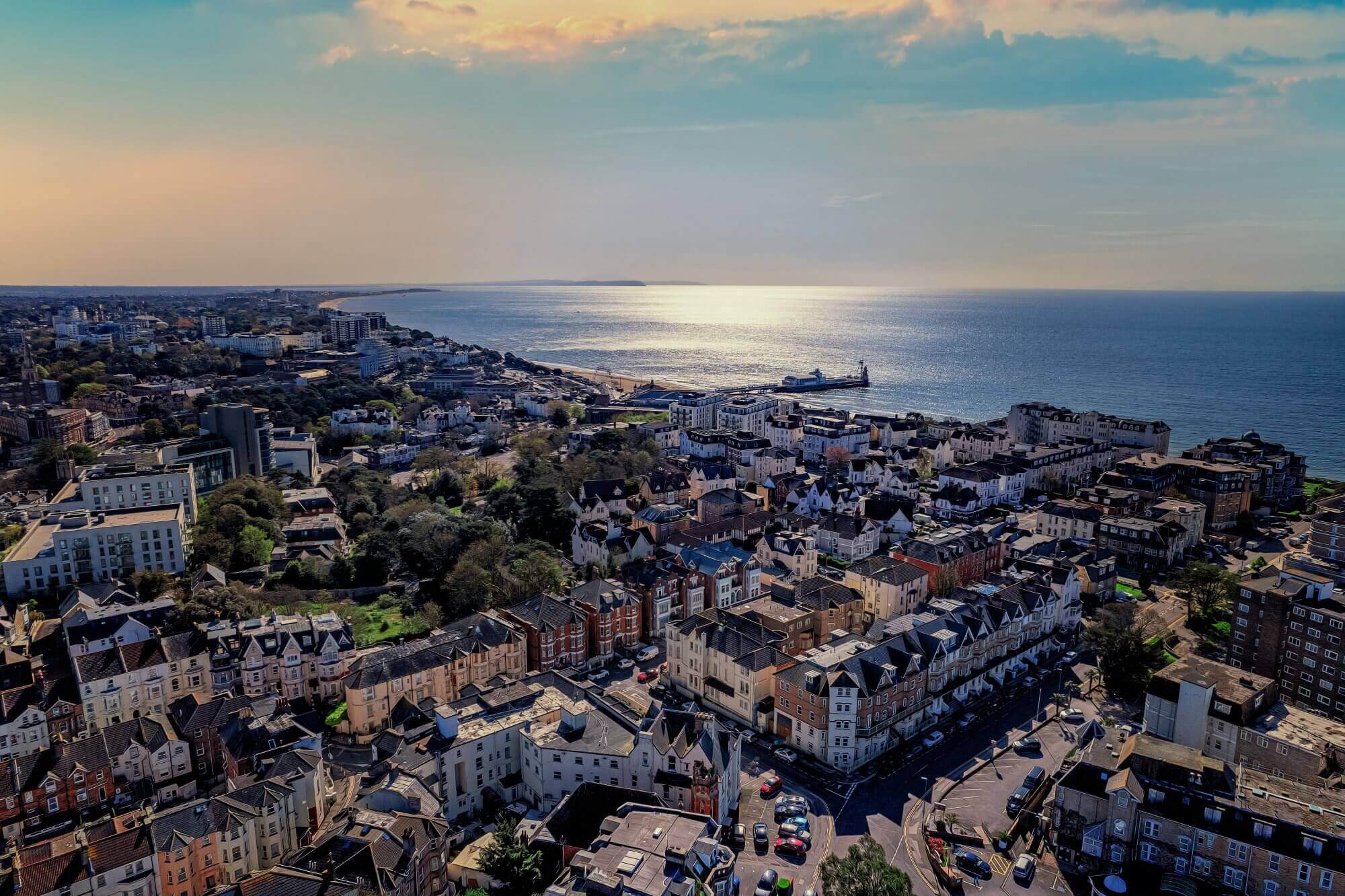 Aerial view of a coastal town with buildings, streets, and the ocean glistening under a setting sun in the background. - Home Instead Bournemouth & Christchurch