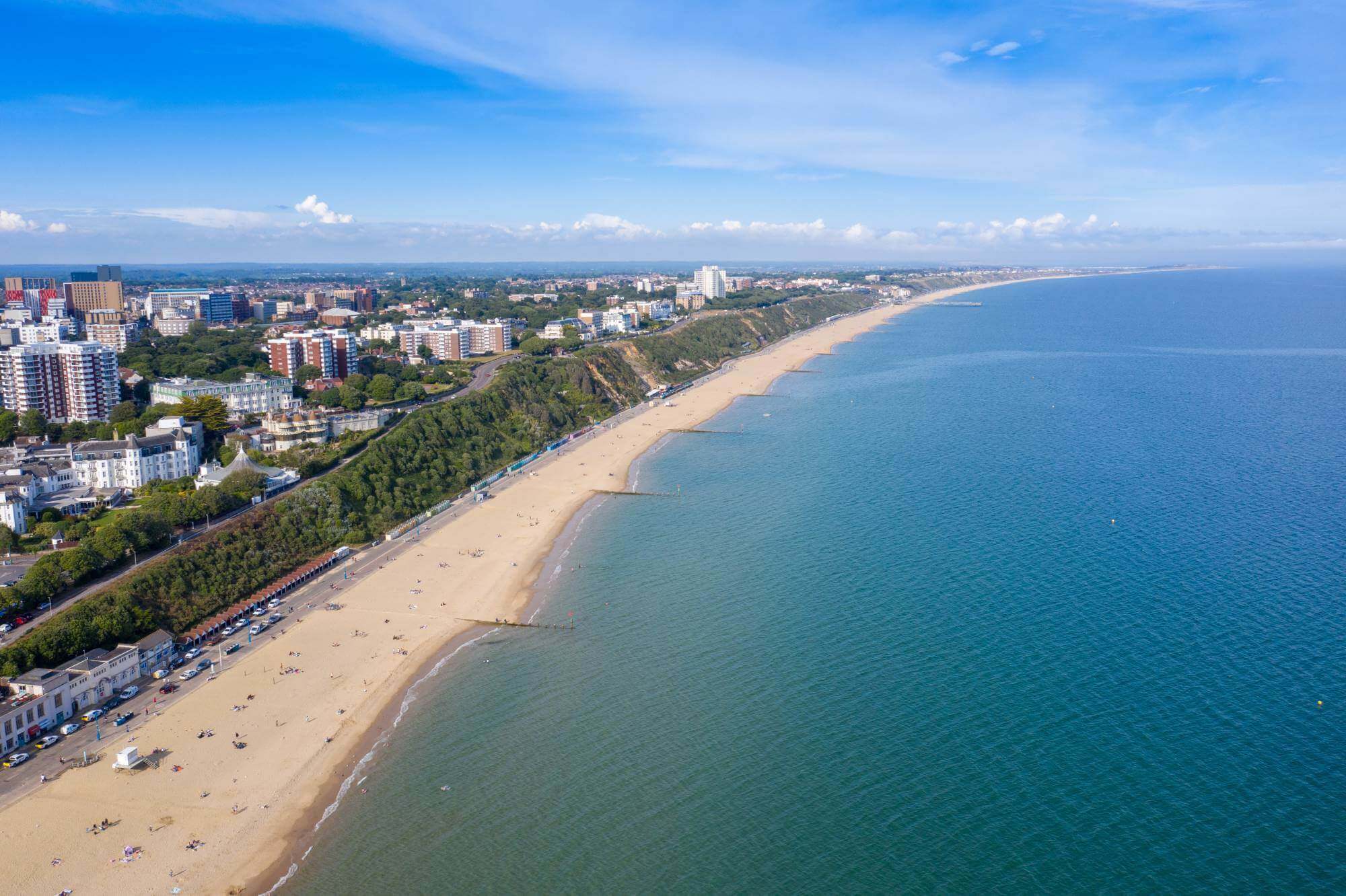 Aerial view of a sandy beach stretching alongside a coastal town with buildings, greenery, and clear blue water under a bright sky. - Home Instead Southampton