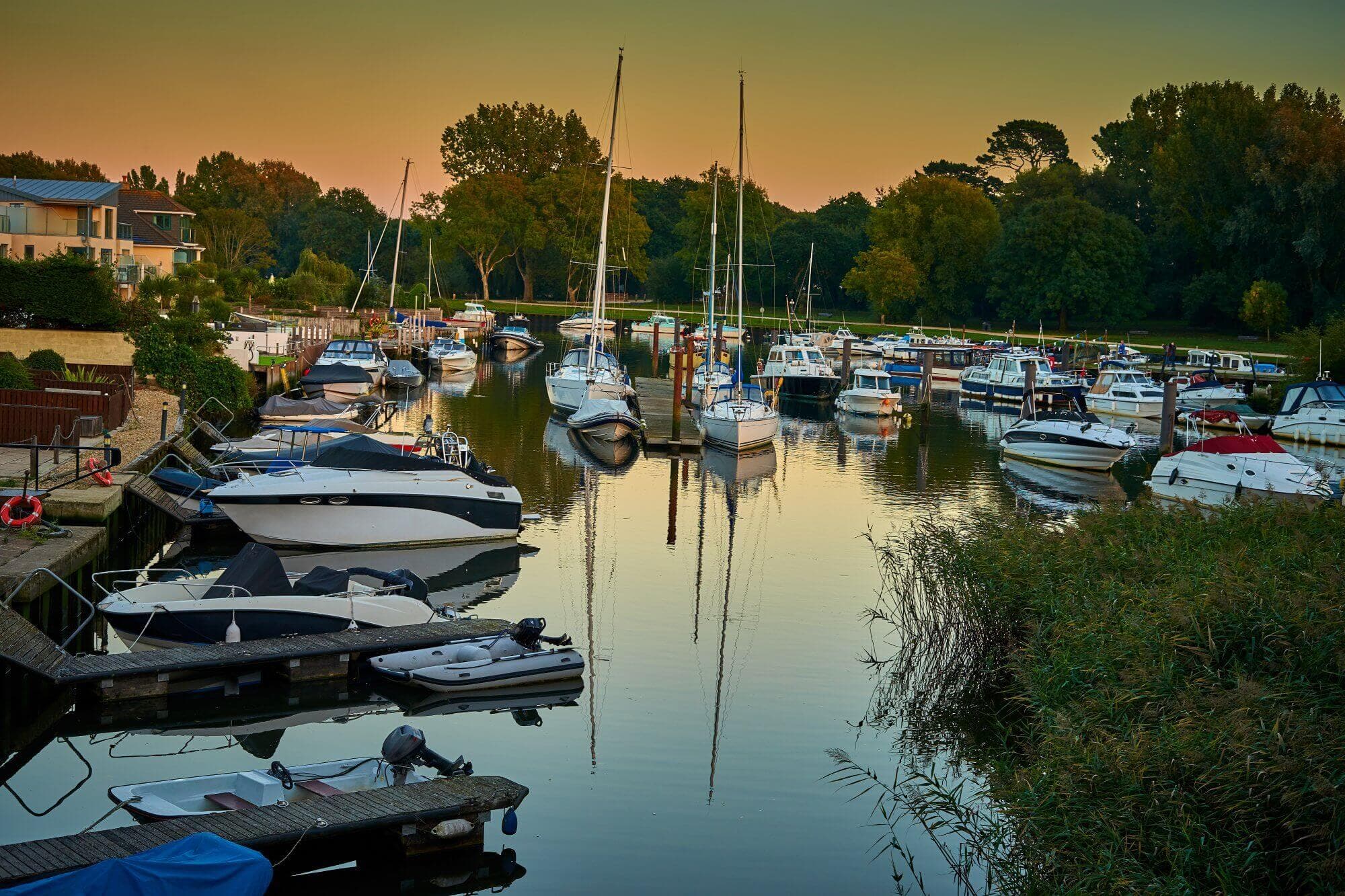 A serene marina at sunset with numerous boats docked, surrounded by lush greenery and buildings on the left. - Home Instead Bournemouth & Christchurch