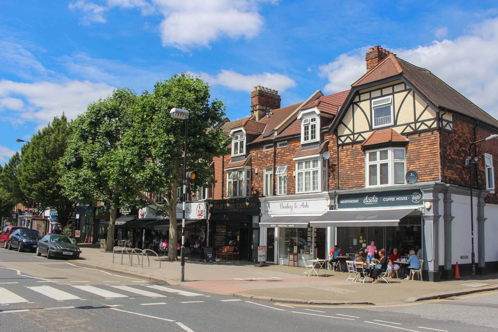 Charming street with people dining outdoors, parked cars, and traditional buildings under a blue sky with clouds. - Home Instead