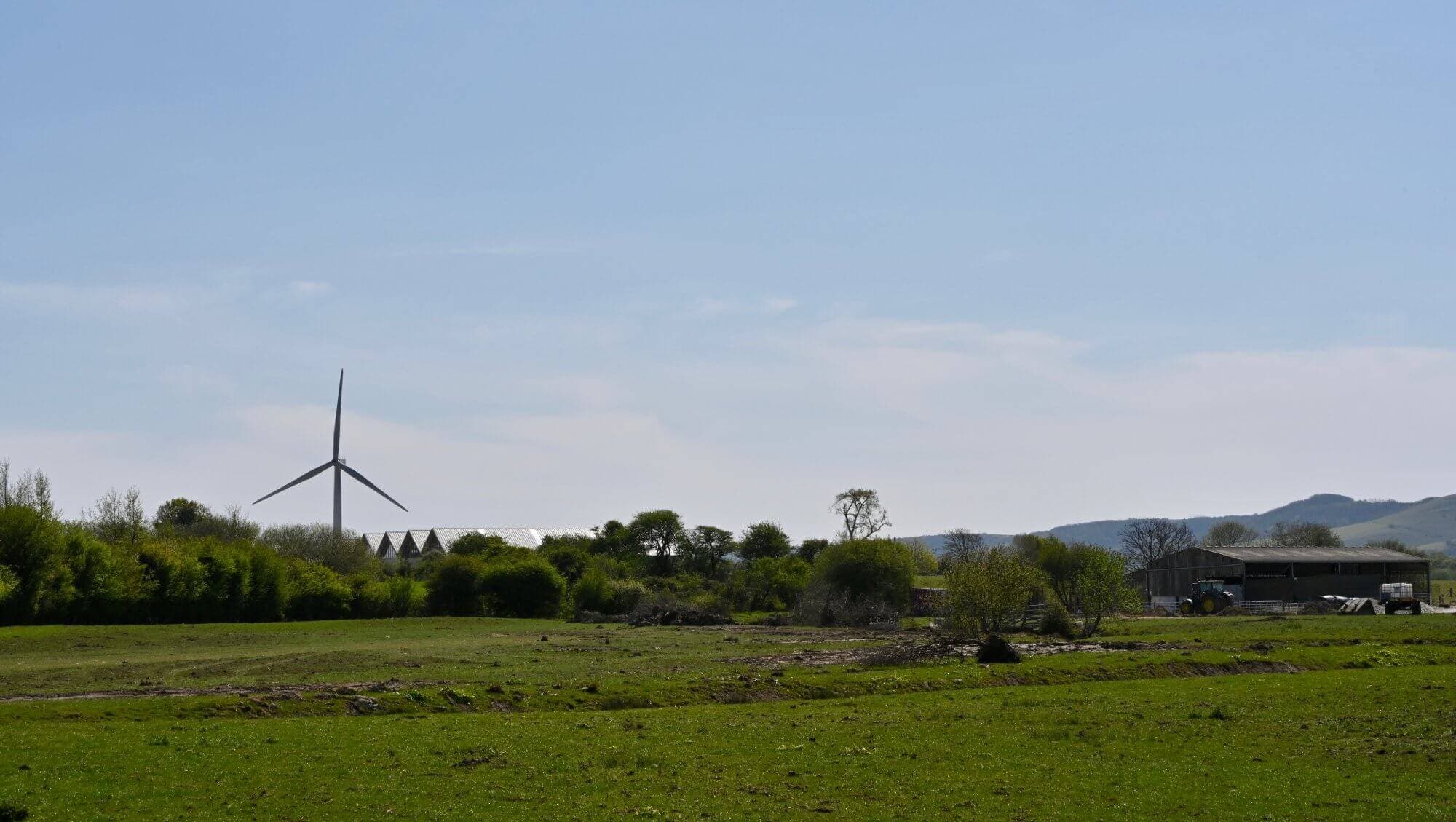 Wind turbine in a rural landscape with green fields, trees, hills, and a small building under a clear blue sky. - Home Instead