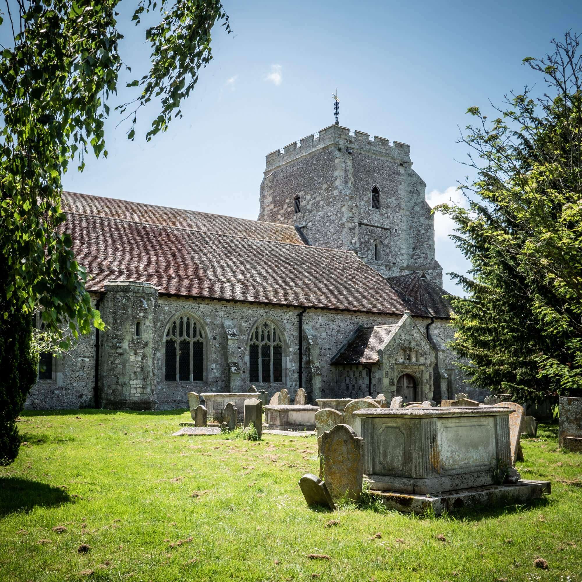 Old stone church with a square tower and gravestones in a sunny, green cemetery. Trees frame the scene. - Home Instead