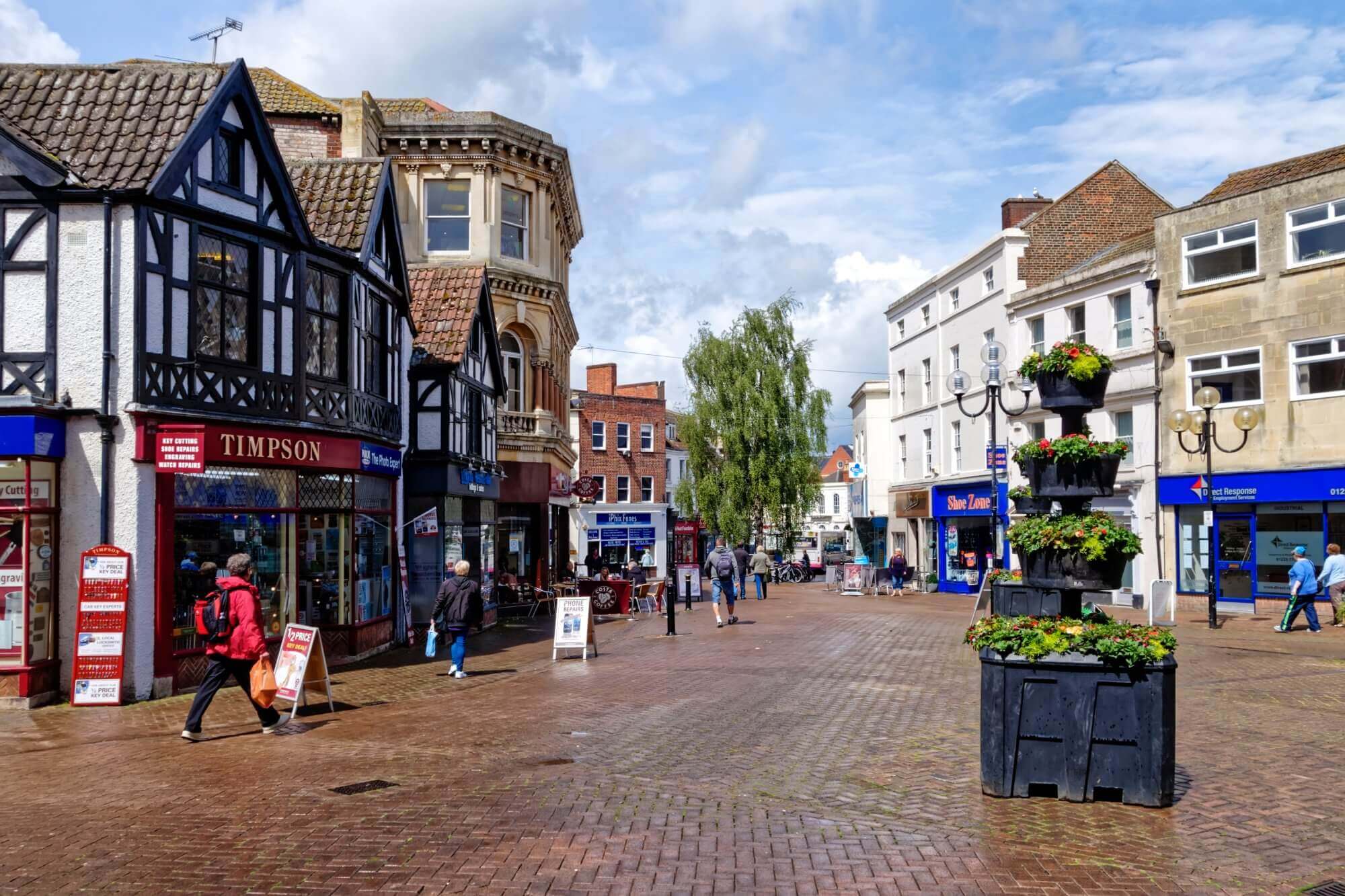 A busy street scene in a town center with shops, a black planter, and people walking around. - Home Instead