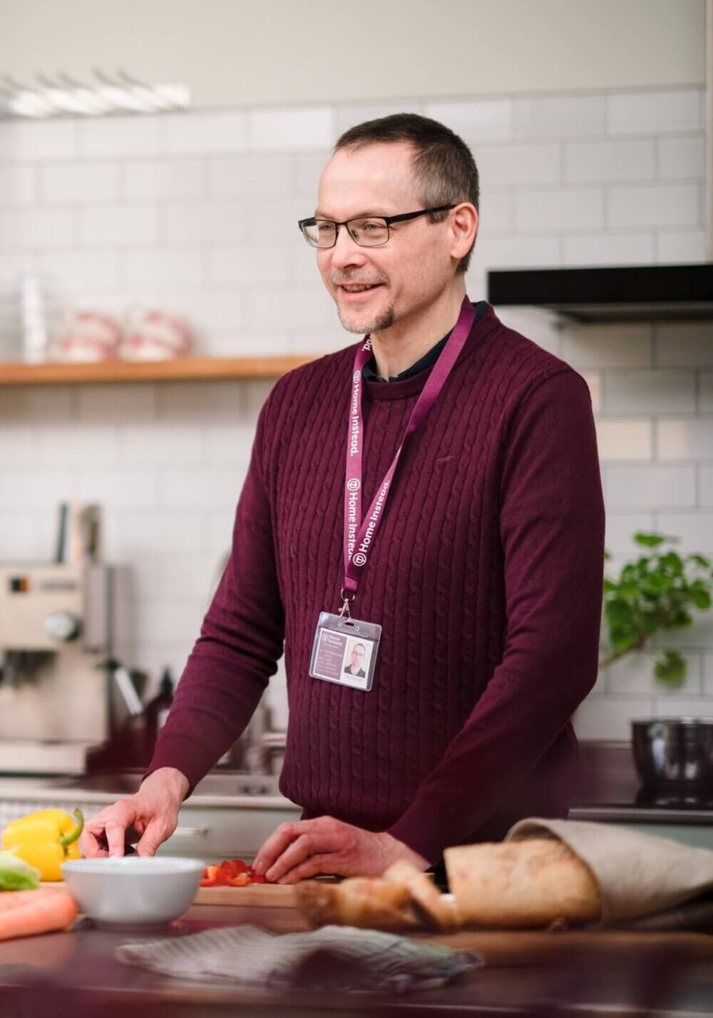 A man in a purple sweater, wearing a nametag, is chopping vegetables in a kitchen with a smile. - Home Instead