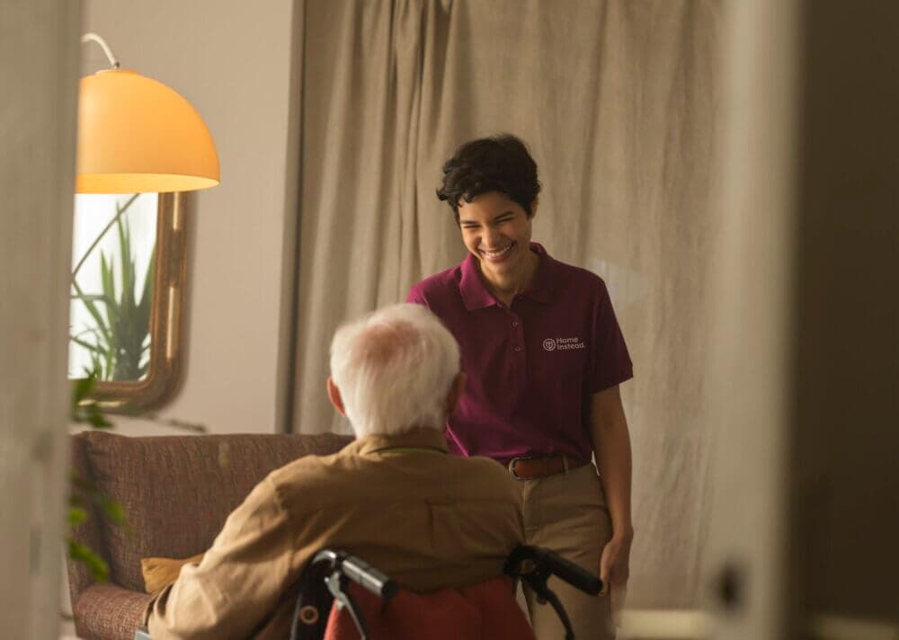 A caregiver smiles warmly at an elderly person in a wheelchair in a cozy, well-lit room. - Home Instead