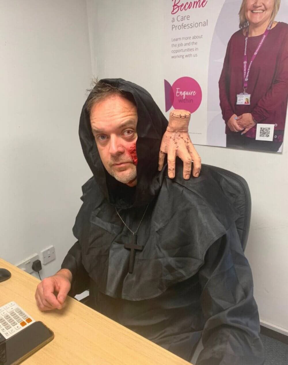 A person in a black hooded costume with a fake hand on their shoulder sits at a desk, with a poster in the background. - Home Instead