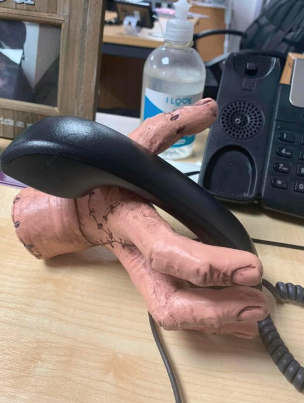 A phone handset held by a realistic, discolored severed hand sculpture on a desk, with office items in the background. - Home Instead