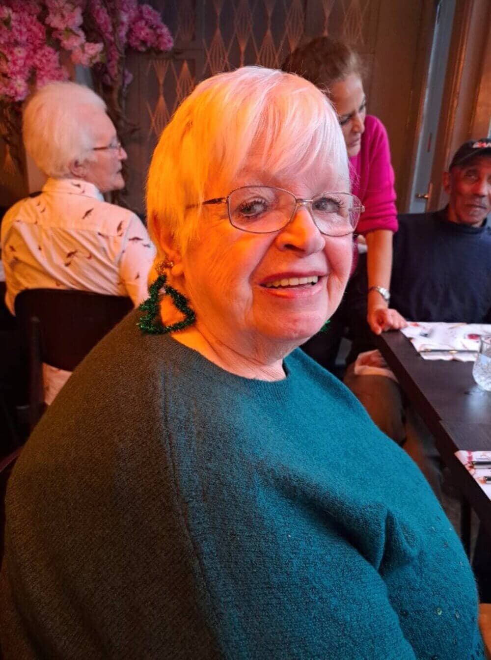 Older woman with glasses and green earrings, smiling at a gathering, with three other people in the background. - Home Instead