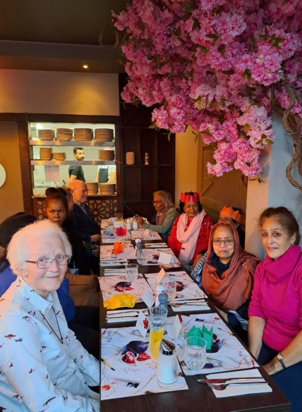 A group of elderly women enjoying a meal together at a restaurant, sitting beneath pink flowering tree decor. - Home Instead