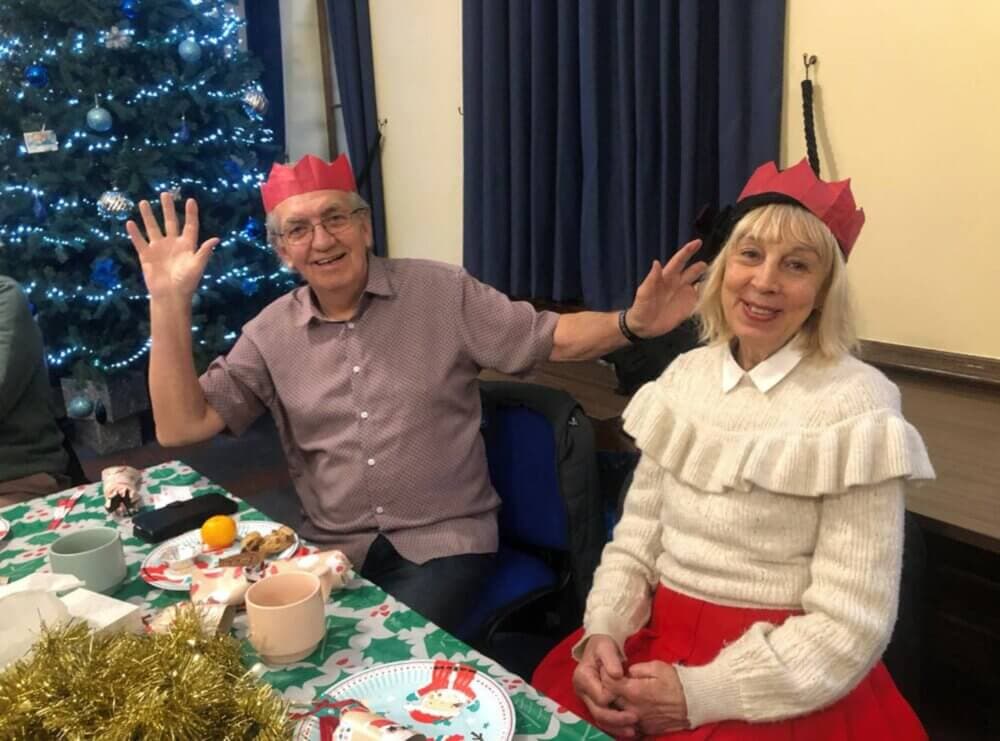 Two people wearing festive hats and sitting at a table beside a decorated Christmas tree, smiling and waving. - Home Instead