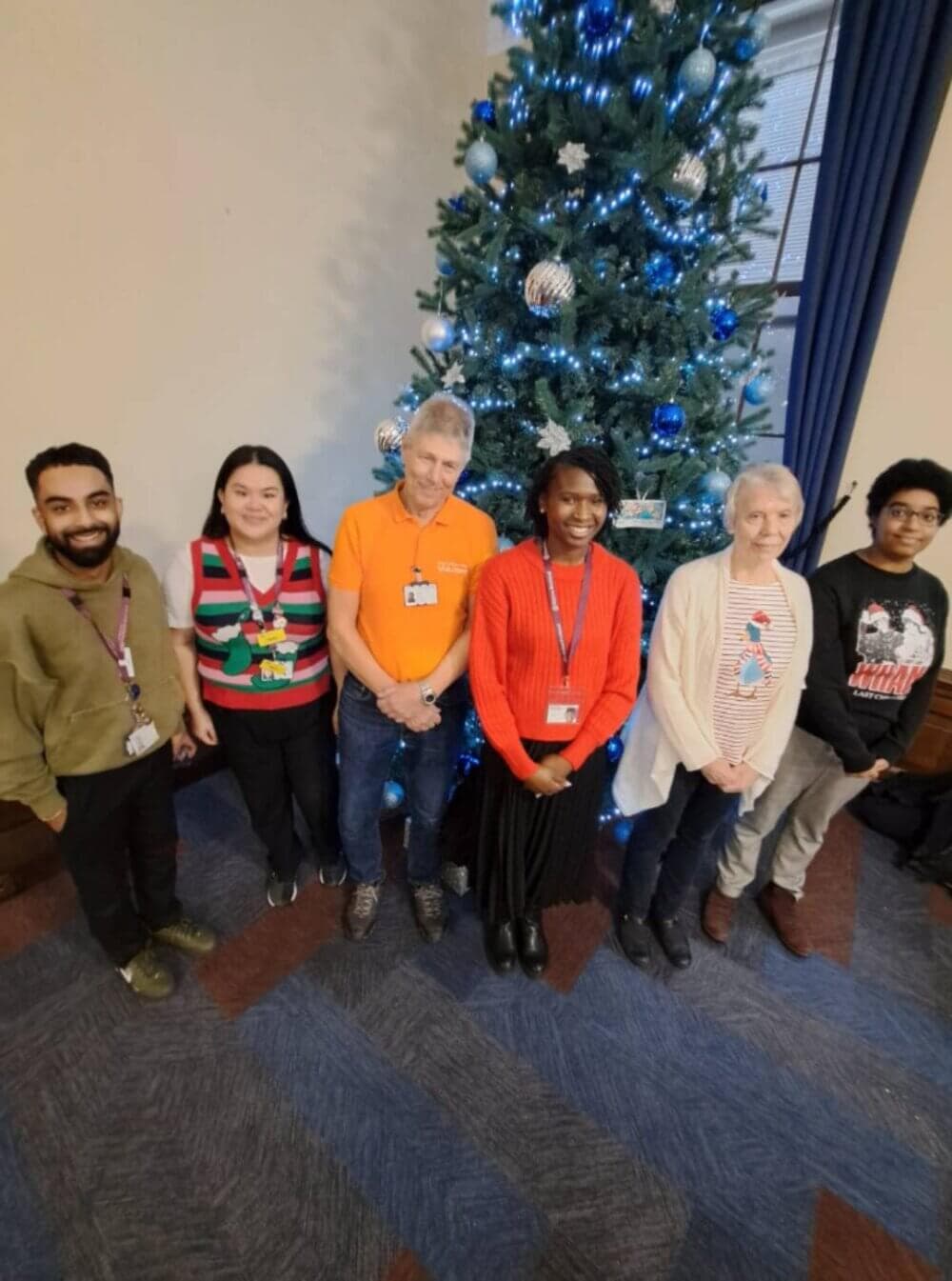 A group of six people standing in front of a decorated Christmas tree in a room with blue patterned carpet. - Home Instead
