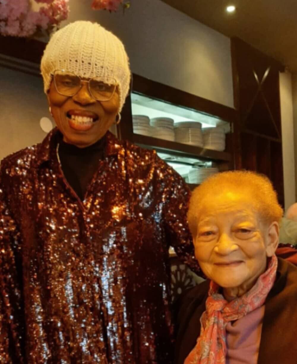 Two elderly women smiling together; one is standing wearing a sequin top and knit cap, the other is seated wearing a scarf. - Home Instead