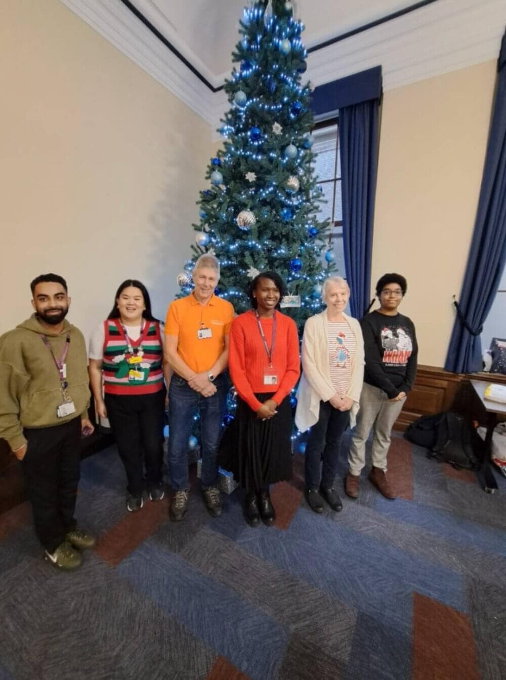 Six people standing in front of a decorated Christmas tree in a room with beige walls and blue curtains. - Home Instead