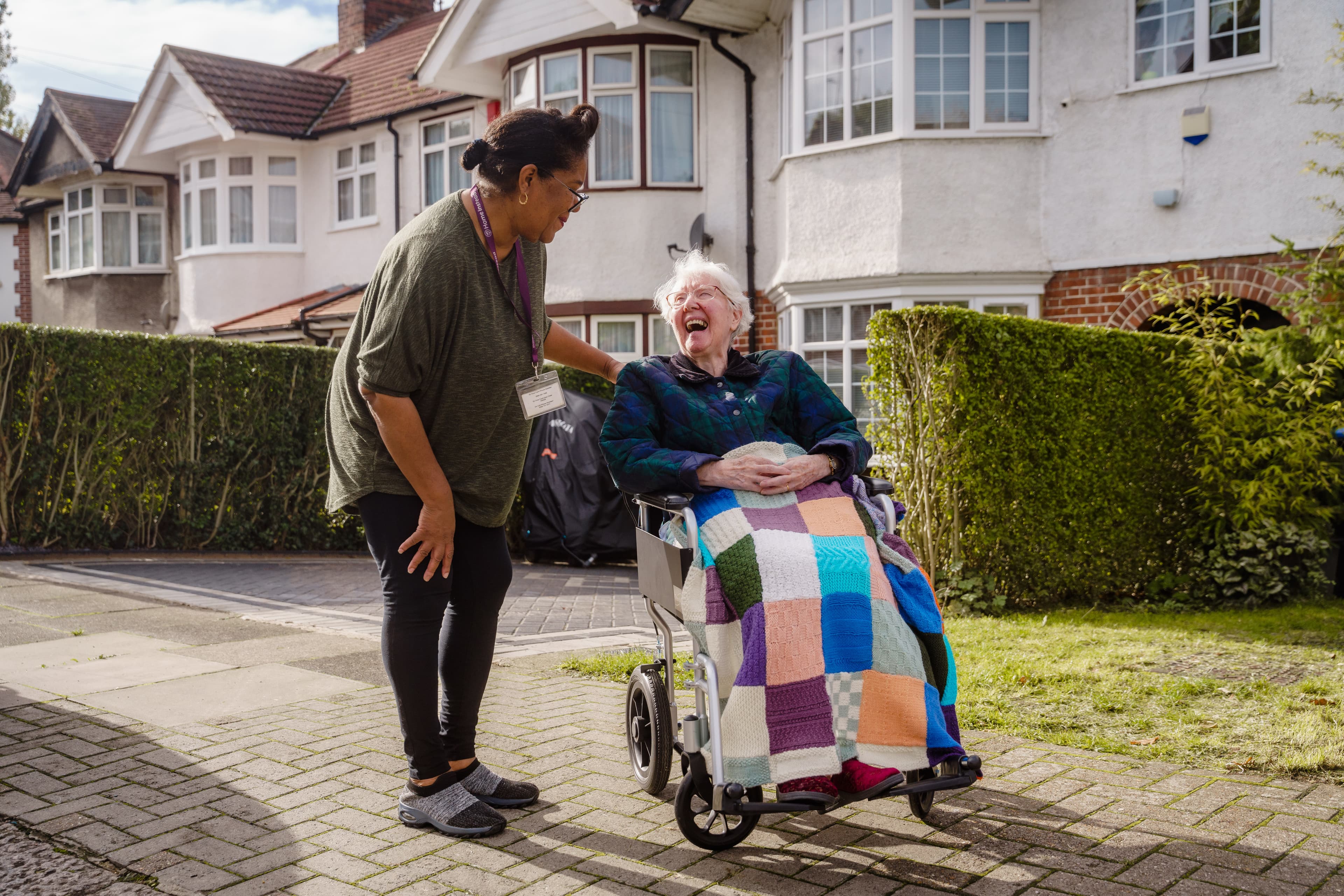 A woman leans to chat with a smiling elderly woman in a wheelchair covered by a colorful quilt, outside a house. - Home Instead