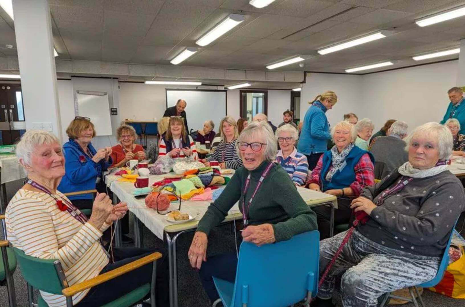 A group of elderly women sit around a table knitting and smiling in a brightly lit room. - Home Instead