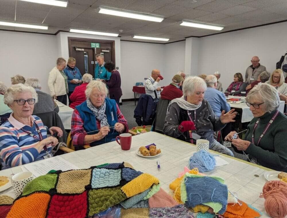 Group of elderly people knitting and socializing at a community center. Yarn, blankets, and snacks are on the tables. - Home Instead