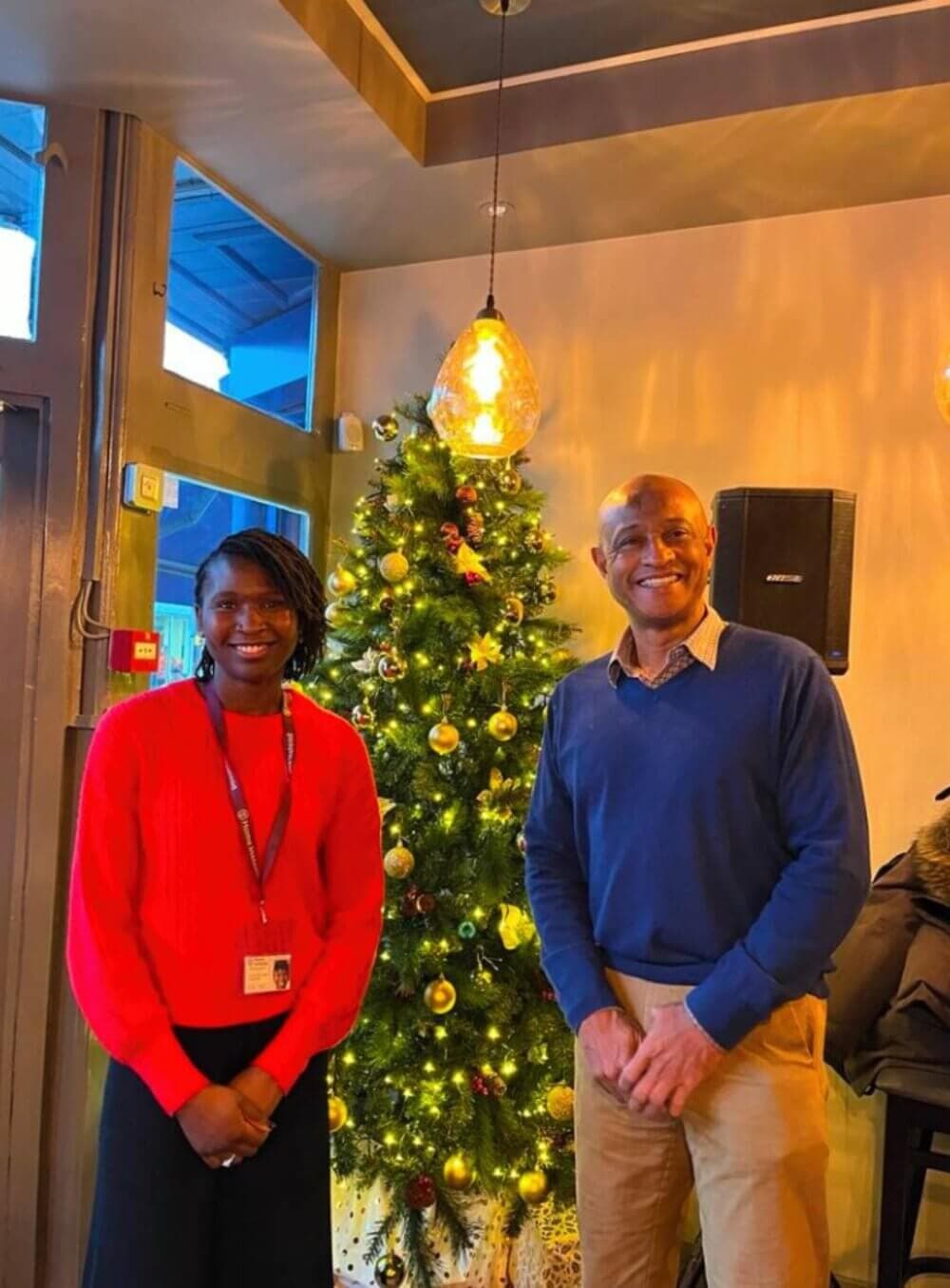 Two people smiling in front of a decorated Christmas tree inside a warmly lit room. - Home Instead