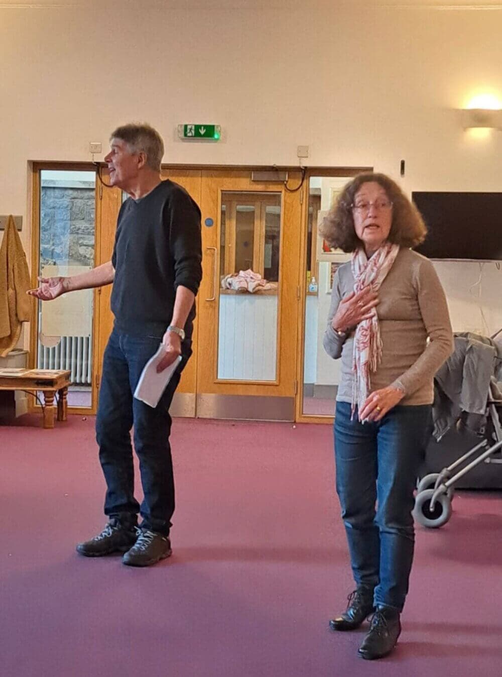 Two people with papers in what seems to be a classroom, performing or rehearsing, with a stroller in the background. - Home Instead