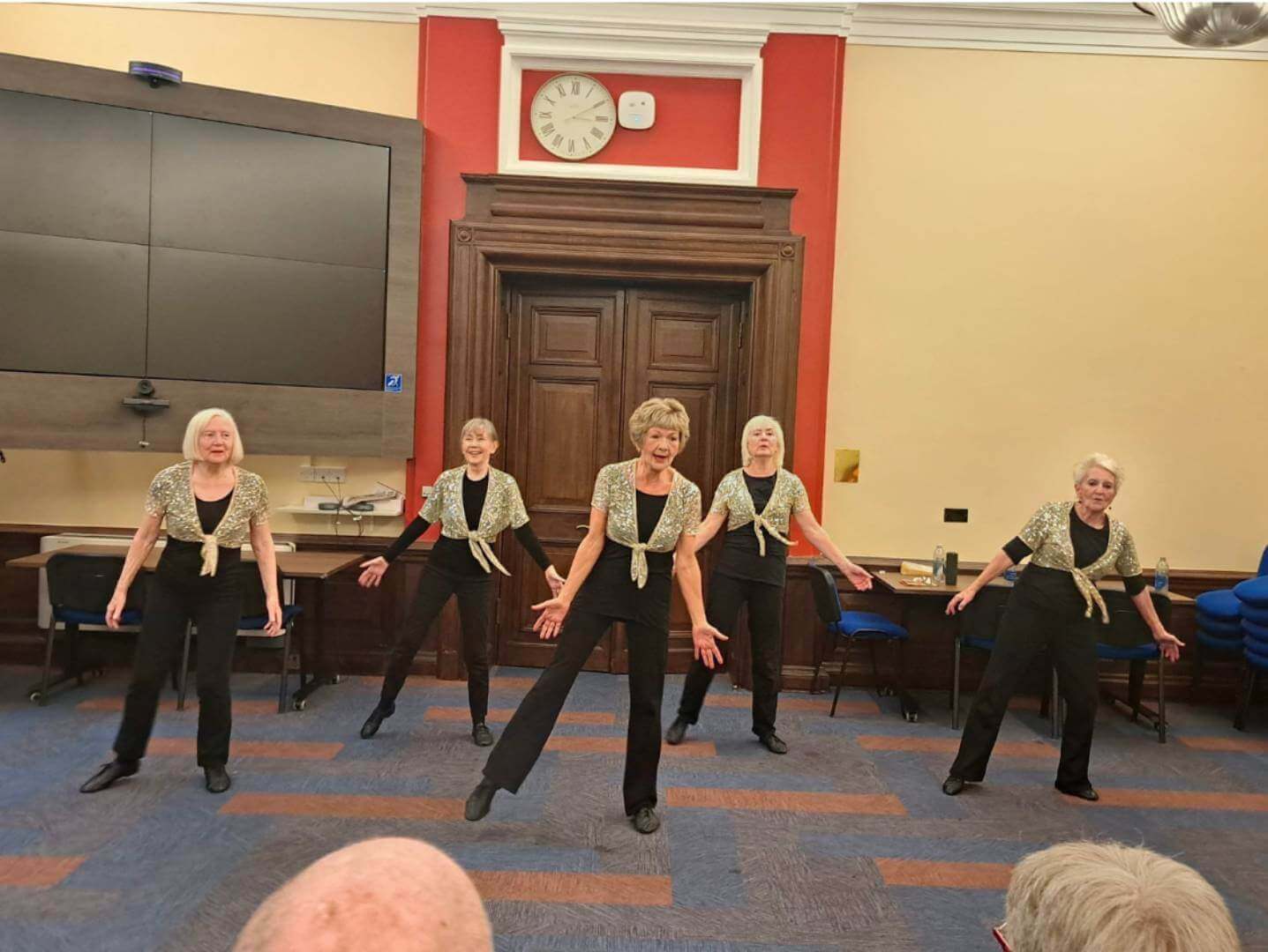 Five women in matching outfits perform a dance routine on a carpeted stage in a wood-paneled room. - Home Instead