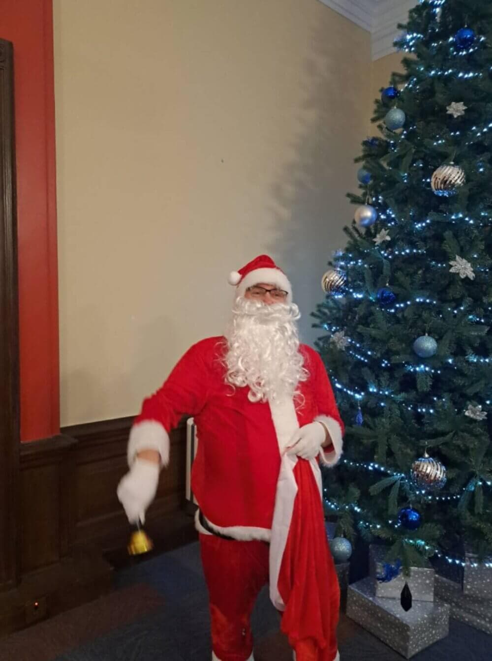 Person dressed as Santa Claus standing next to a decorated Christmas tree, holding a bell and a red sack. - Home Instead