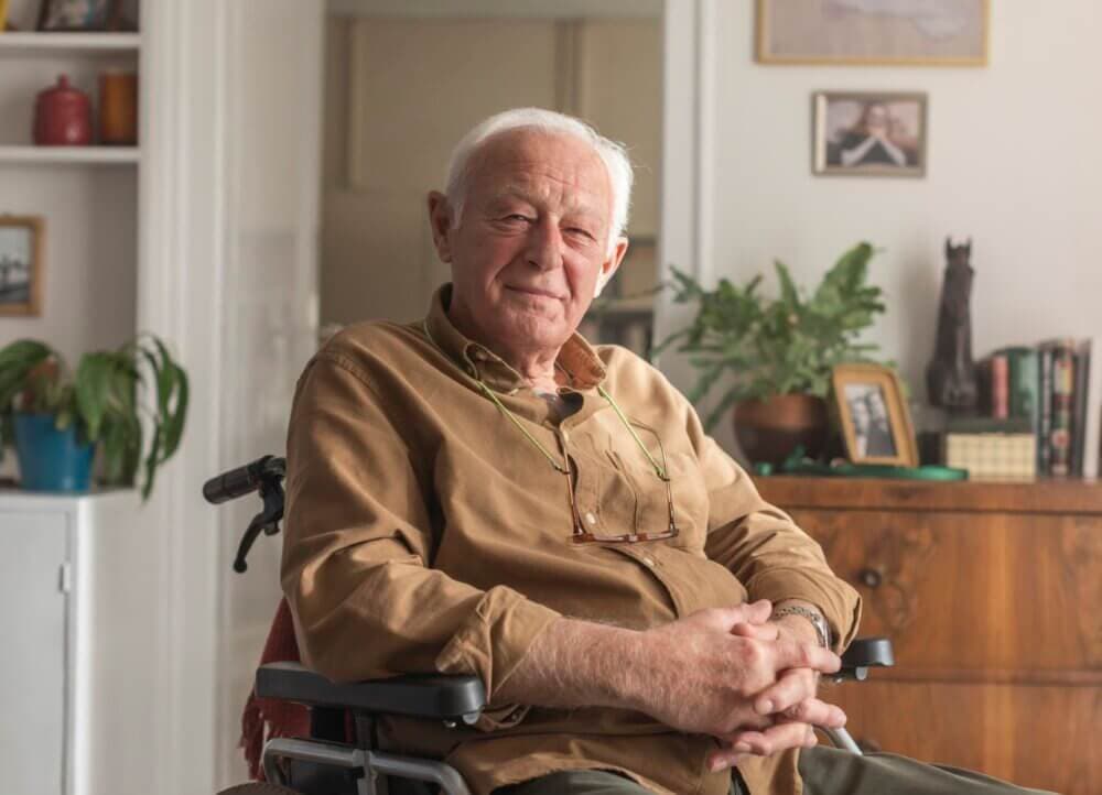 Elderly man sitting in a wheelchair, smiling, with a cozy, decorated room in the background. - Home Instead