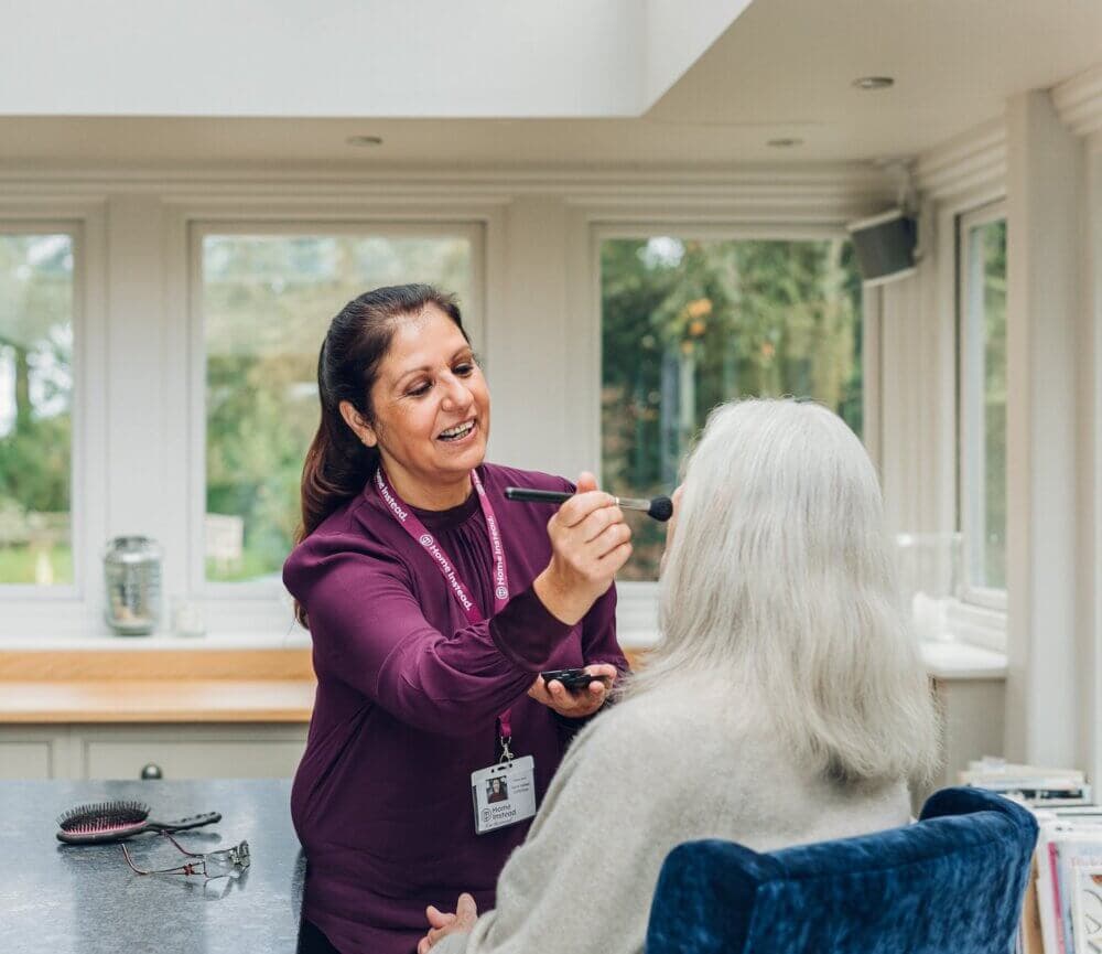 A woman is applying makeup to an elderly woman seated in a brightly lit room with large windows. - Home Instead