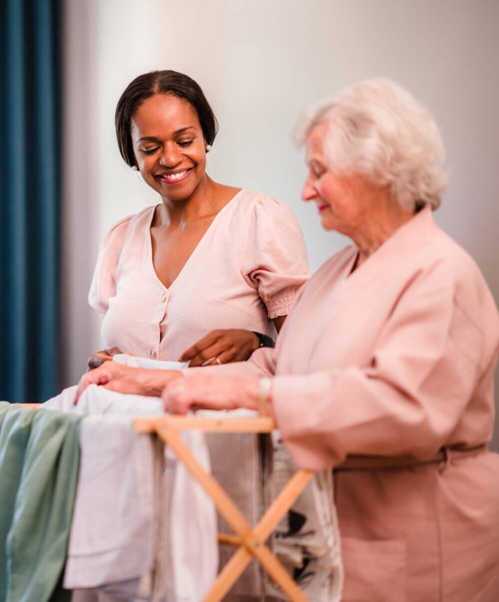A young woman and an elderly woman smile while folding laundry together, with a clothes drying rack in front of them. - Home Instead