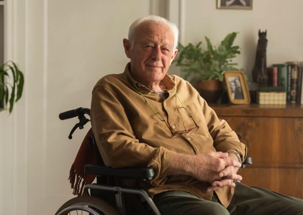 An elderly man in a wheelchair, wearing a brown shirt, poses indoors with a small smile, surrounded by plants and books. - Home Instead