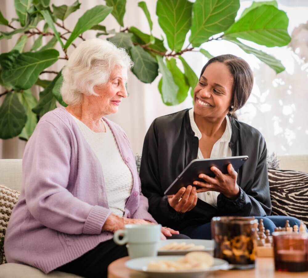A smiling young woman shows a tablet to an elderly woman on a cozy couch with a large plant in the background. - Home Instead