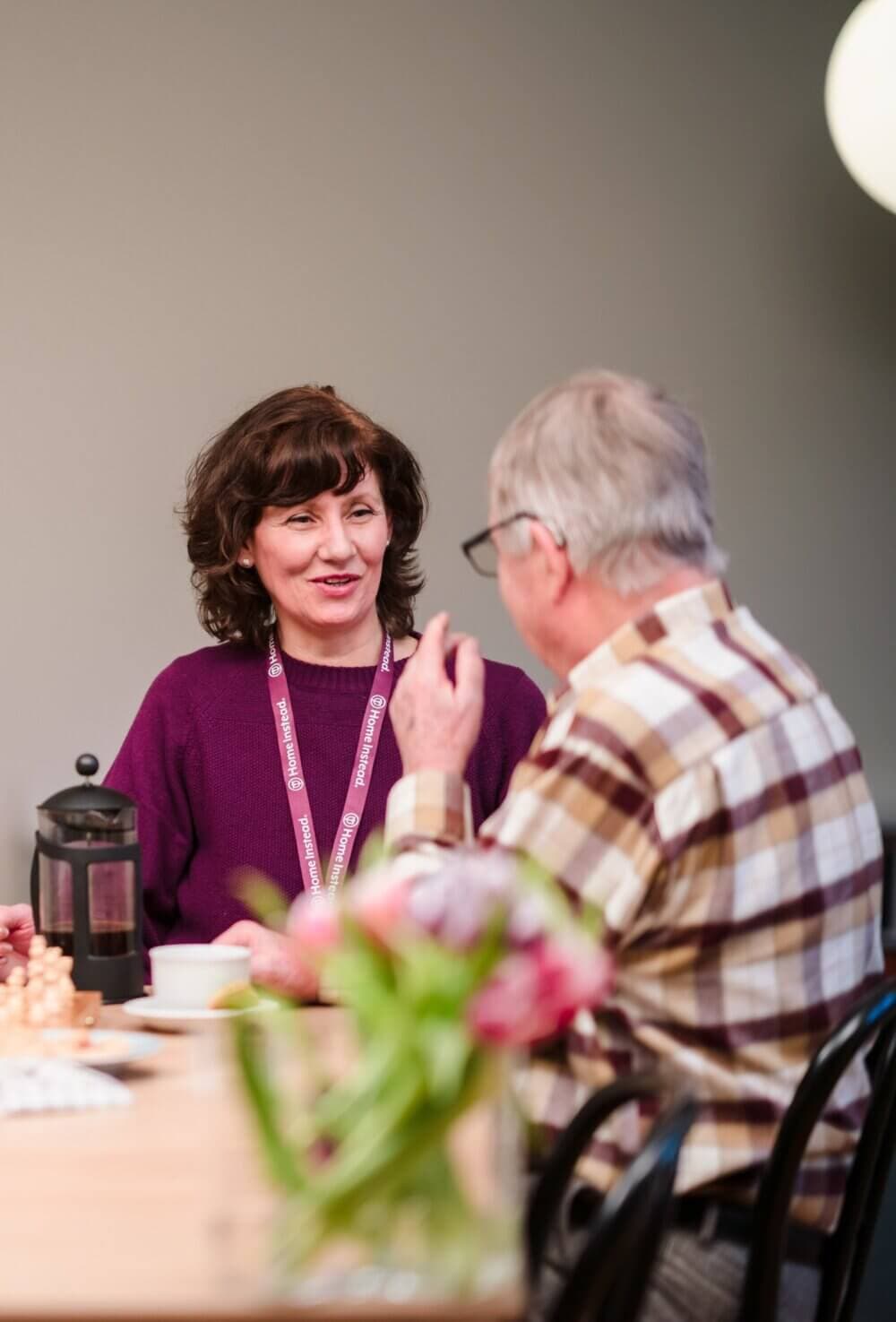 Two people conversing at a table with a French press, coffee cups, and flowers in the foreground. - Home Instead