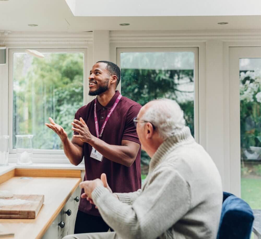 A caregiver in a maroon shirt and an elderly man in a sweater are conversing and smiling in a bright, airy room. - Home Instead