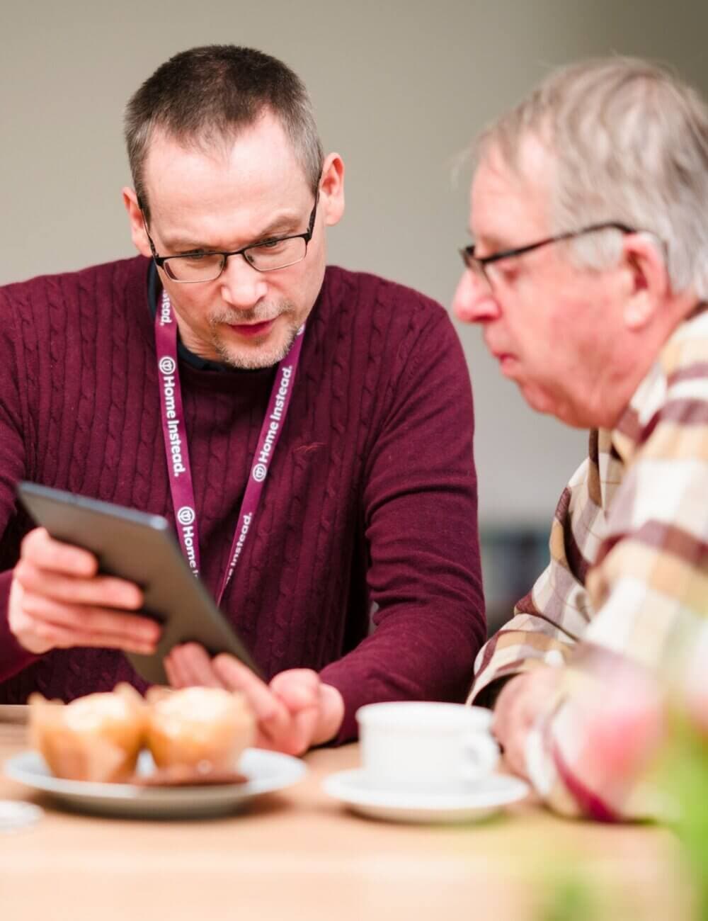 A man helps an older man use a tablet at a table with muffins and a coffee mug. - Home Instead