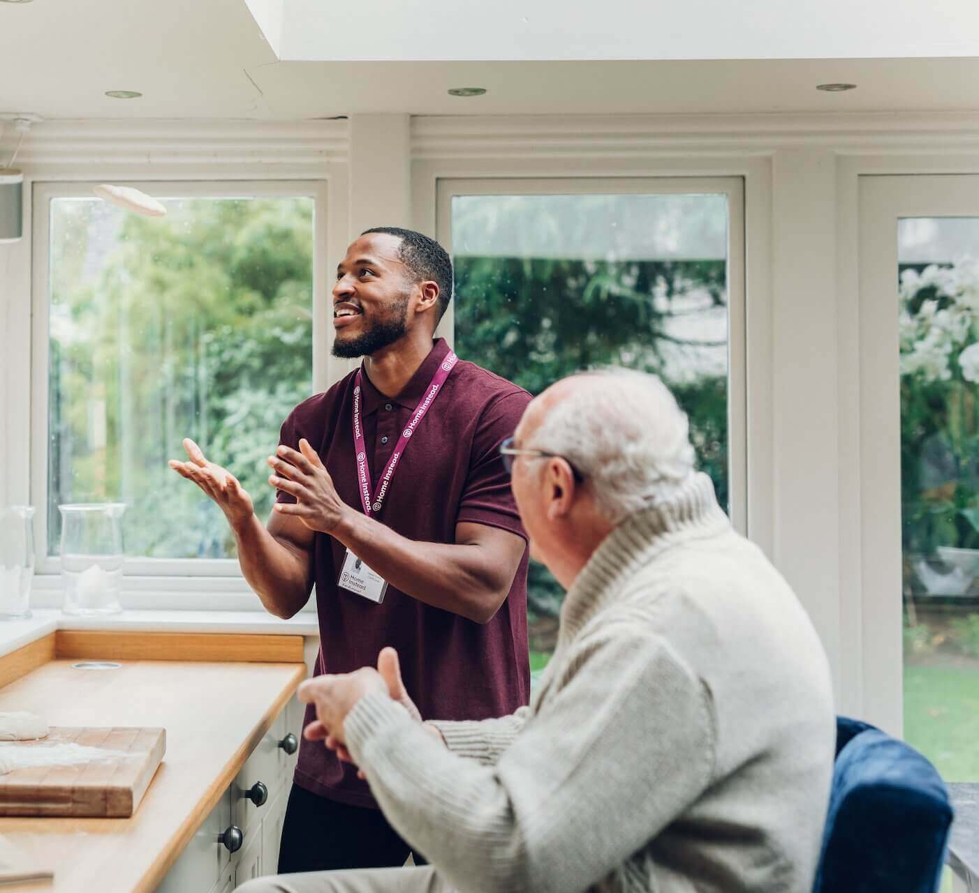 A caregiver in a maroon shirt and an elderly man in a sweater are conversing and smiling in a bright, airy room. - Home Instead