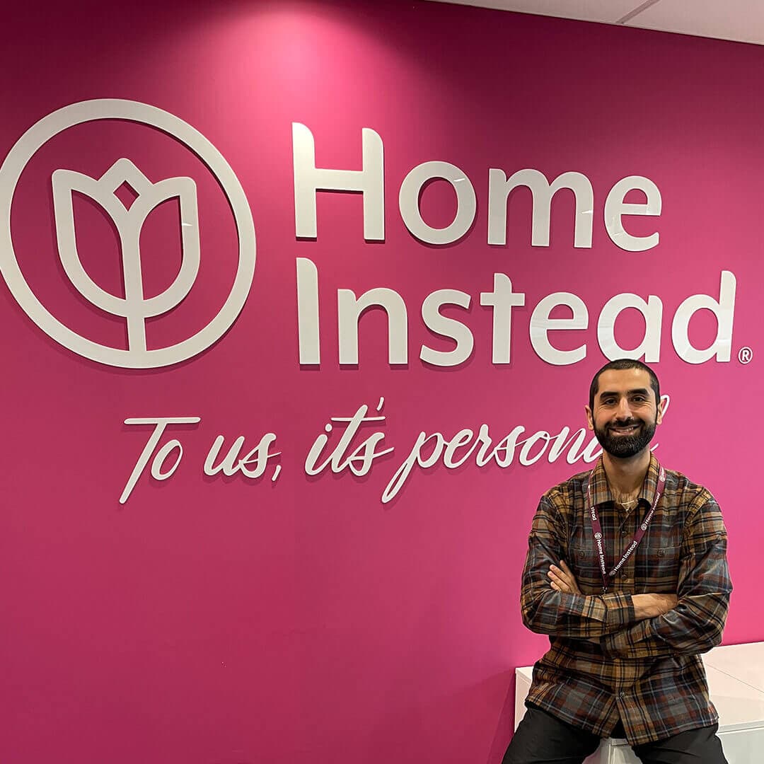 A man with crossed arms stands in front of a pink wall with the Home Instead logo and the slogan "To us, it's personal. - Home Instead