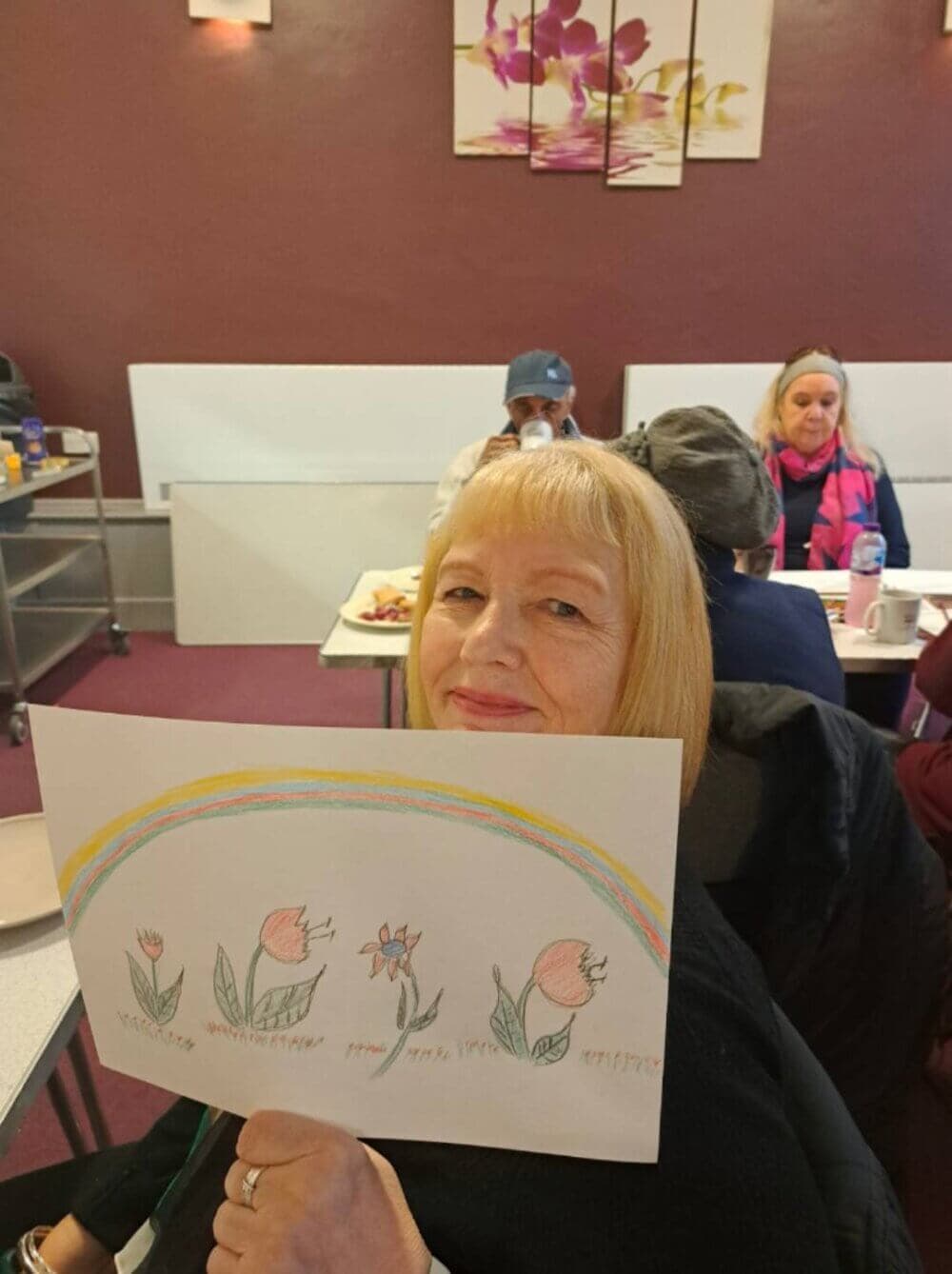 A woman holds a drawing of flowers and a rainbow in a room with people seated at tables in the background. - Home Instead