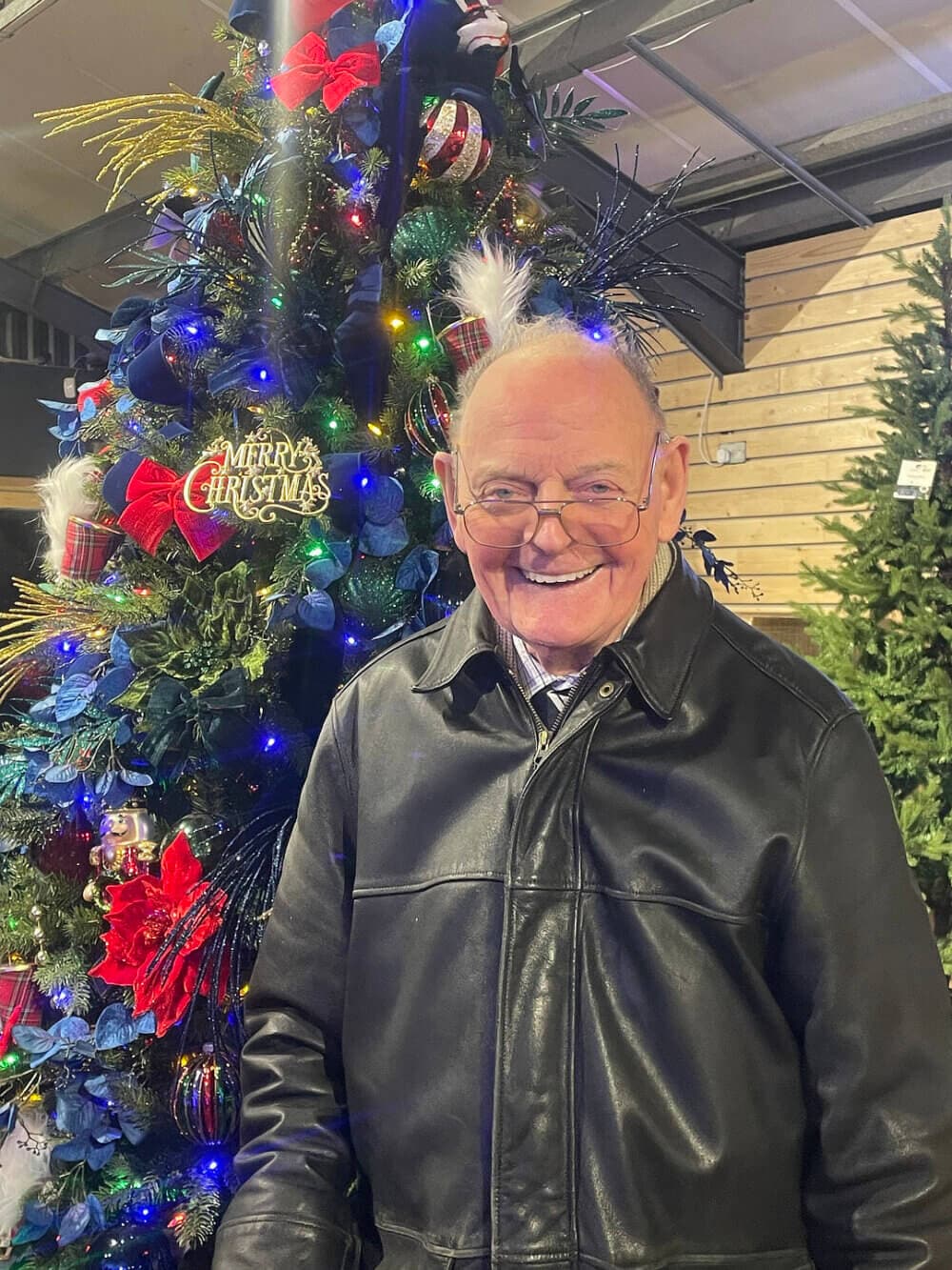 An elderly man in a black leather jacket stands smiling in front of a decorated Christmas tree. - Home Instead