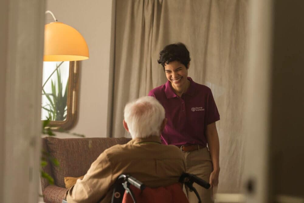 A caregiver in a maroon uniform smiling and talking to an elderly person in a wheelchair in a cozy room. - Home Instead