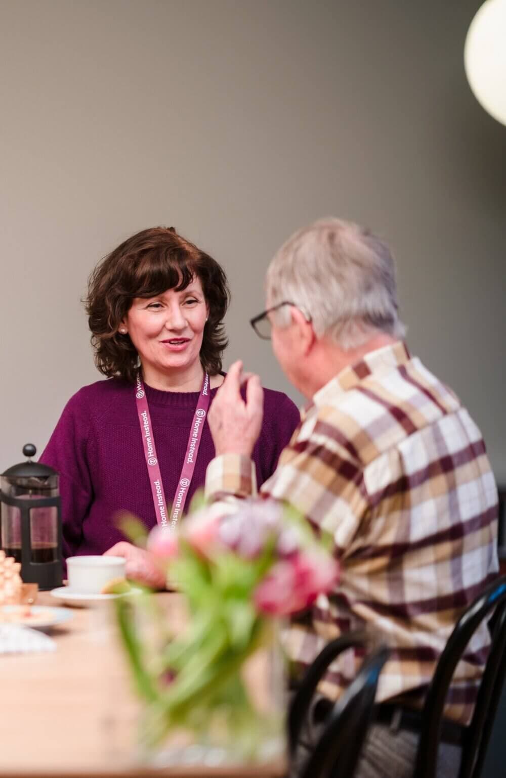 A woman and an elderly man sitting at a table, smiling and looking at a tablet together. - Home Instead