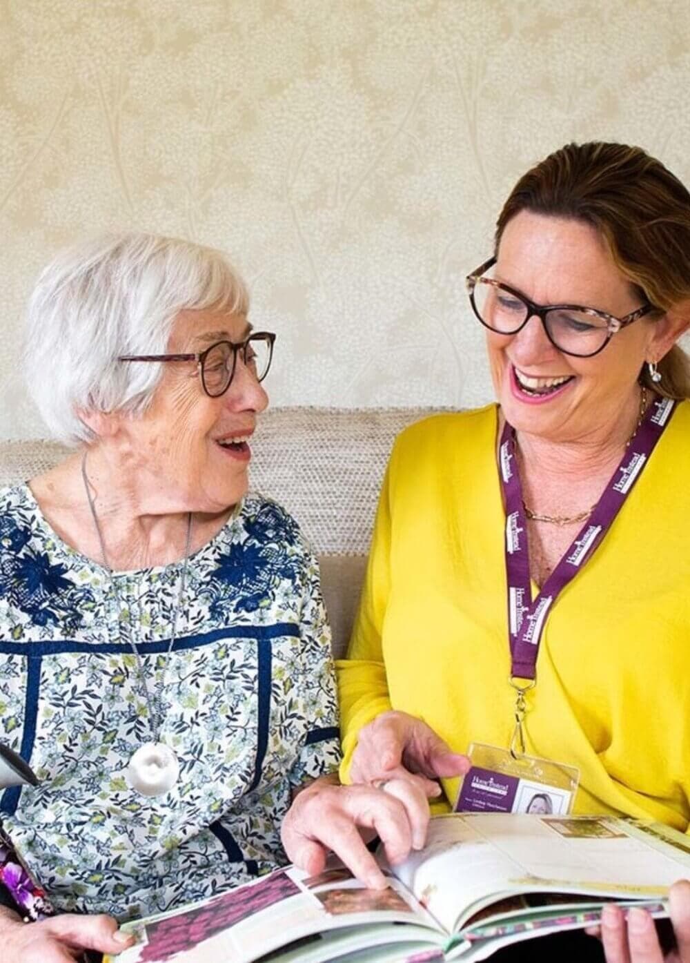 An elderly woman and a caregiver sit together, both smiling and looking at an open book. - Home Instead