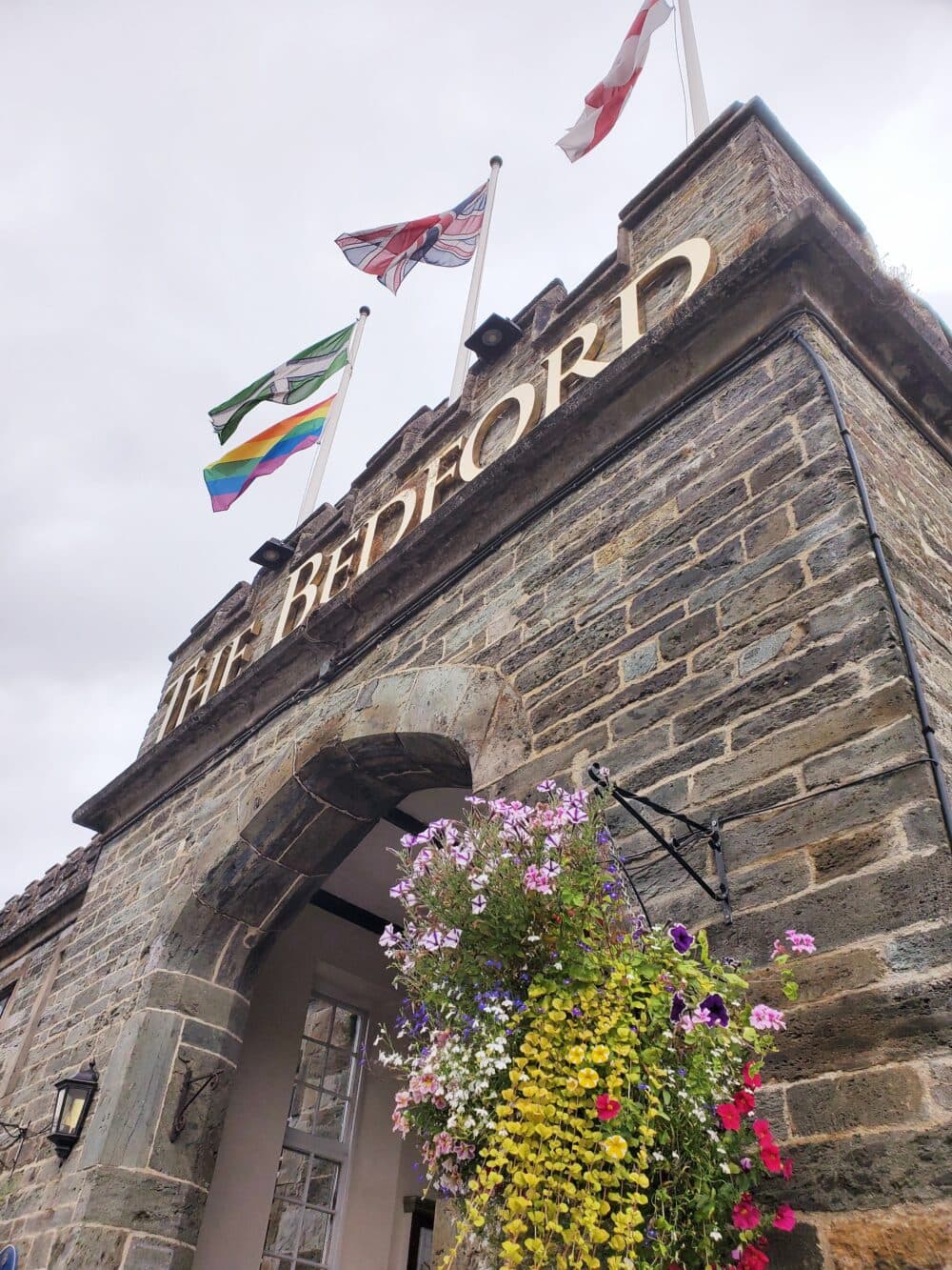 Stone entrance with flags and flowers under "The Bedford" sign, against a cloudy sky. - Home Instead
