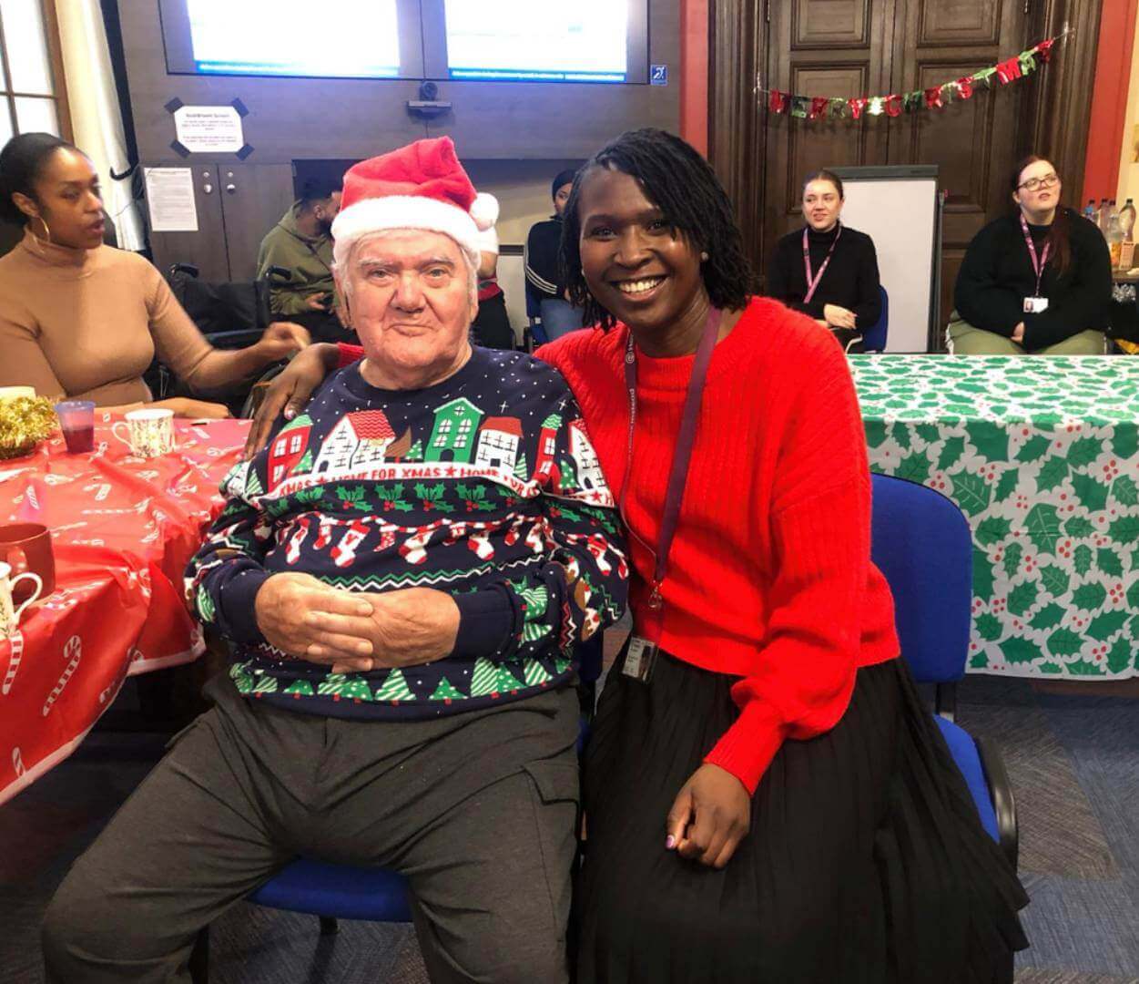 Two people, one wearing a Santa hat, smile together at a festive gathering with holiday decorations in the background. - Home Instead