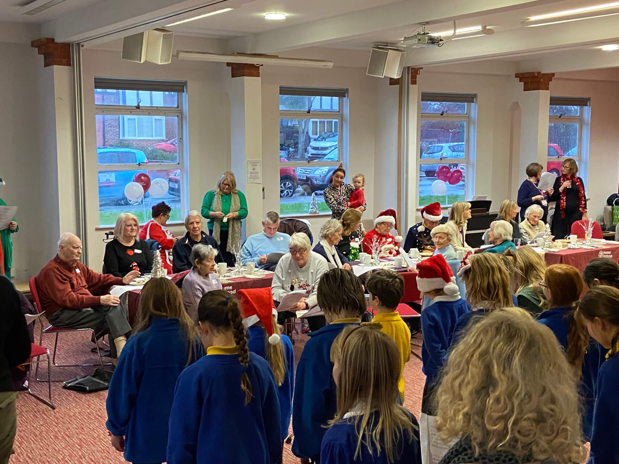 A group of children singing to elderly people at a Christmas gathering inside a community room. - Home Instead