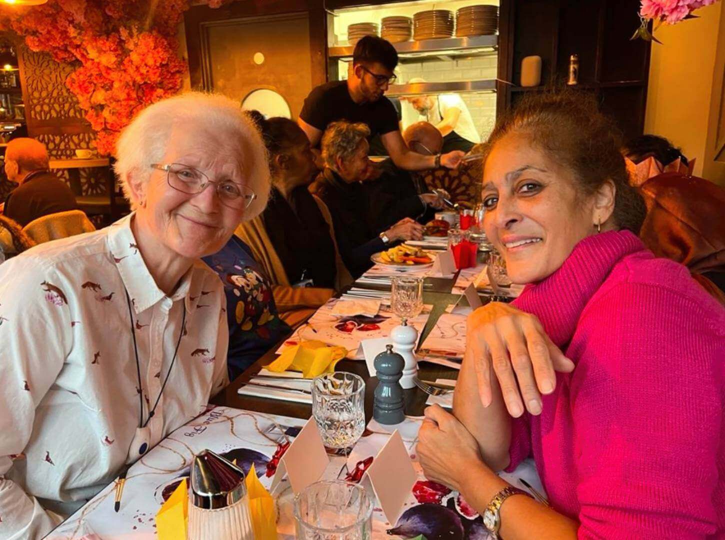 Two elderly women smiling at a decorated restaurant table with other patrons in the background. - Home Instead