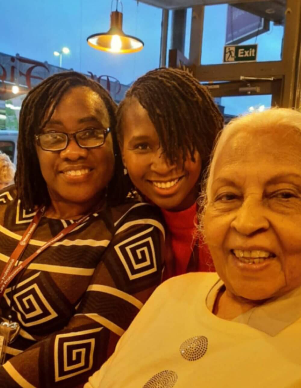 Three women smiling together at a restaurant, warmly lit with pendant lights and an exit sign visible in the background. - Home Instead
