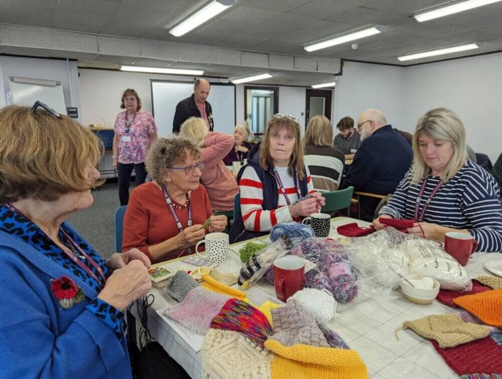 A group of people sitting around a table knitting and chatting in a well-lit room with yarn and knitted items on the table. - Home Instead
