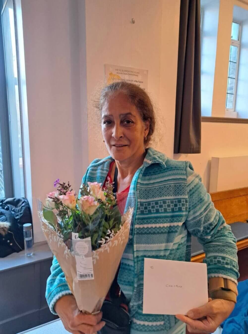 A woman in a blue patterned jacket holds a bouquet of flowers and a white envelope while smiling. - Home Instead
