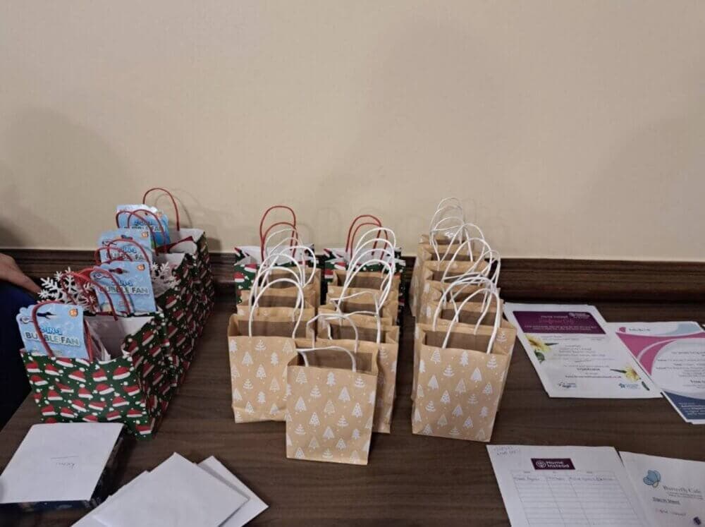 Gift bags with holiday designs and white handles arranged on a table, alongside paperwork and envelopes. - Home Instead