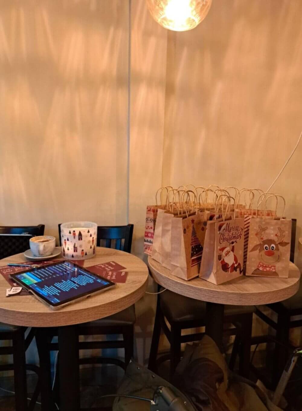 Two tables with Christmas-themed gift bags, a tablet, and a cup inside a warmly lit room. - Home Instead